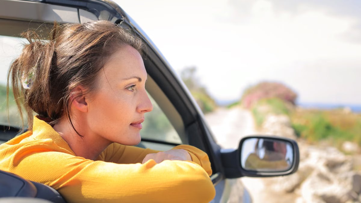 Profile view of a woman leaning out a convertible car enjoying a sunny day on a scenic road.